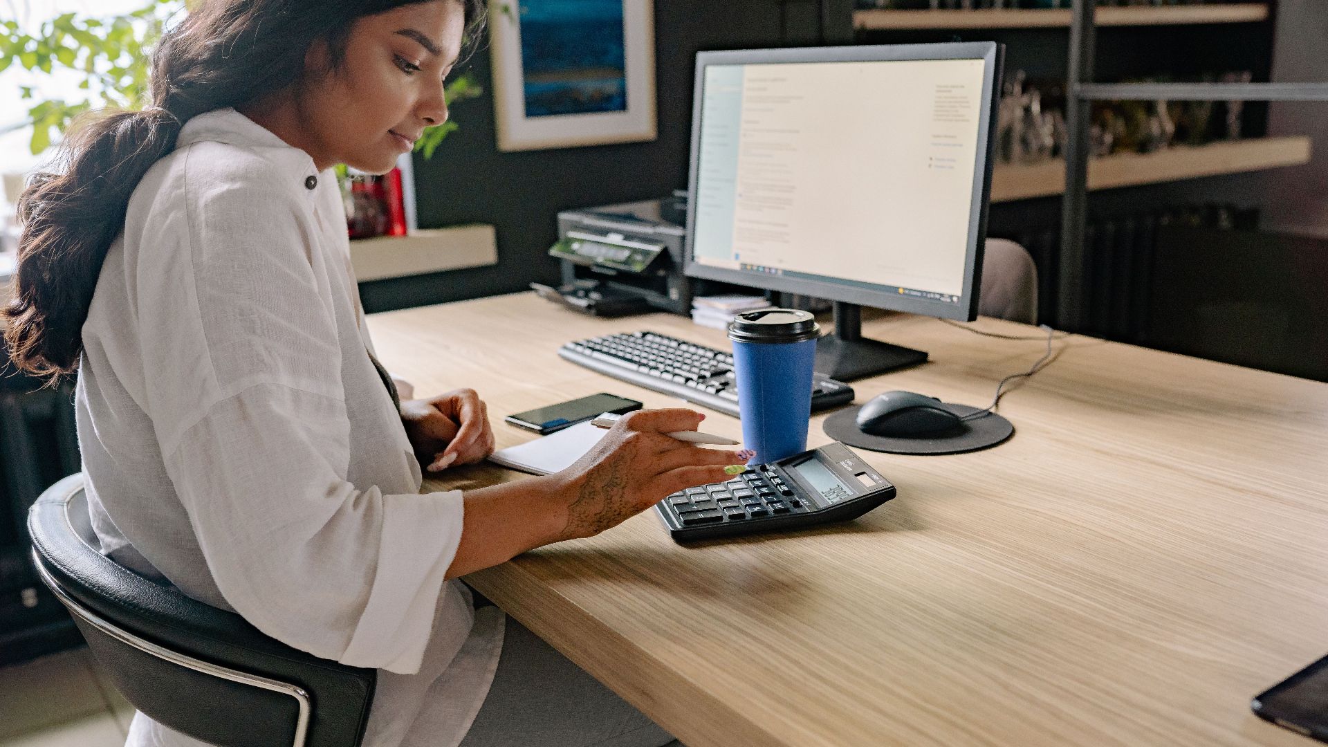 A professional woman using a calculator and computer at an office desk, focused on her work.