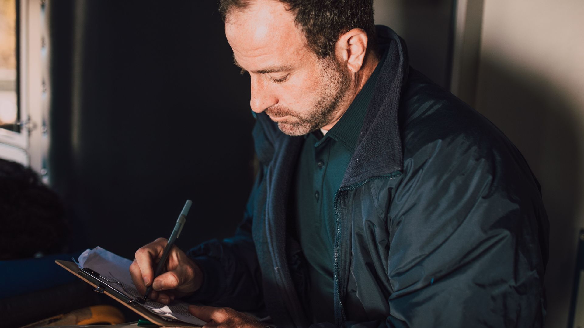 Adult male writing notes on a clipboard inside. Concentrated and thoughtful atmosphere.