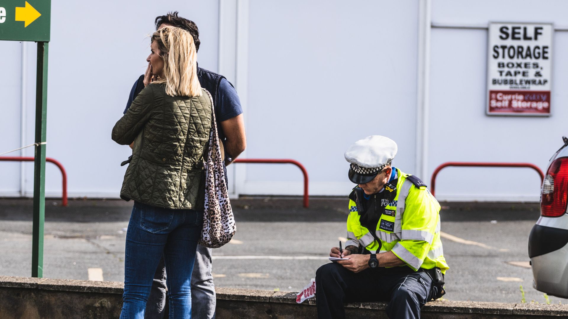 Police officer writing a ticket near a self-storage area with people in the background.