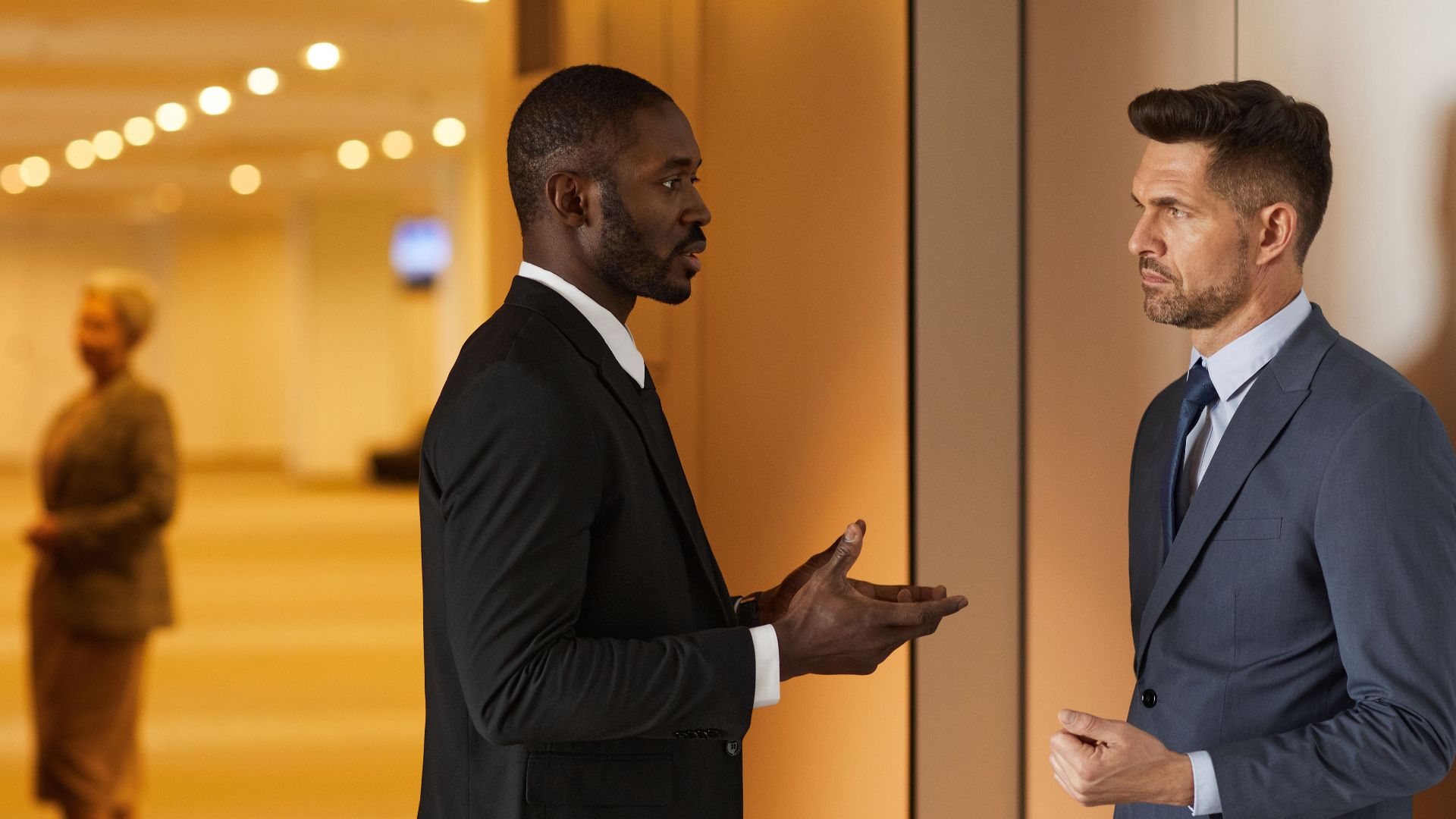 Two men in suits having a discussion in a modern hallway setting.