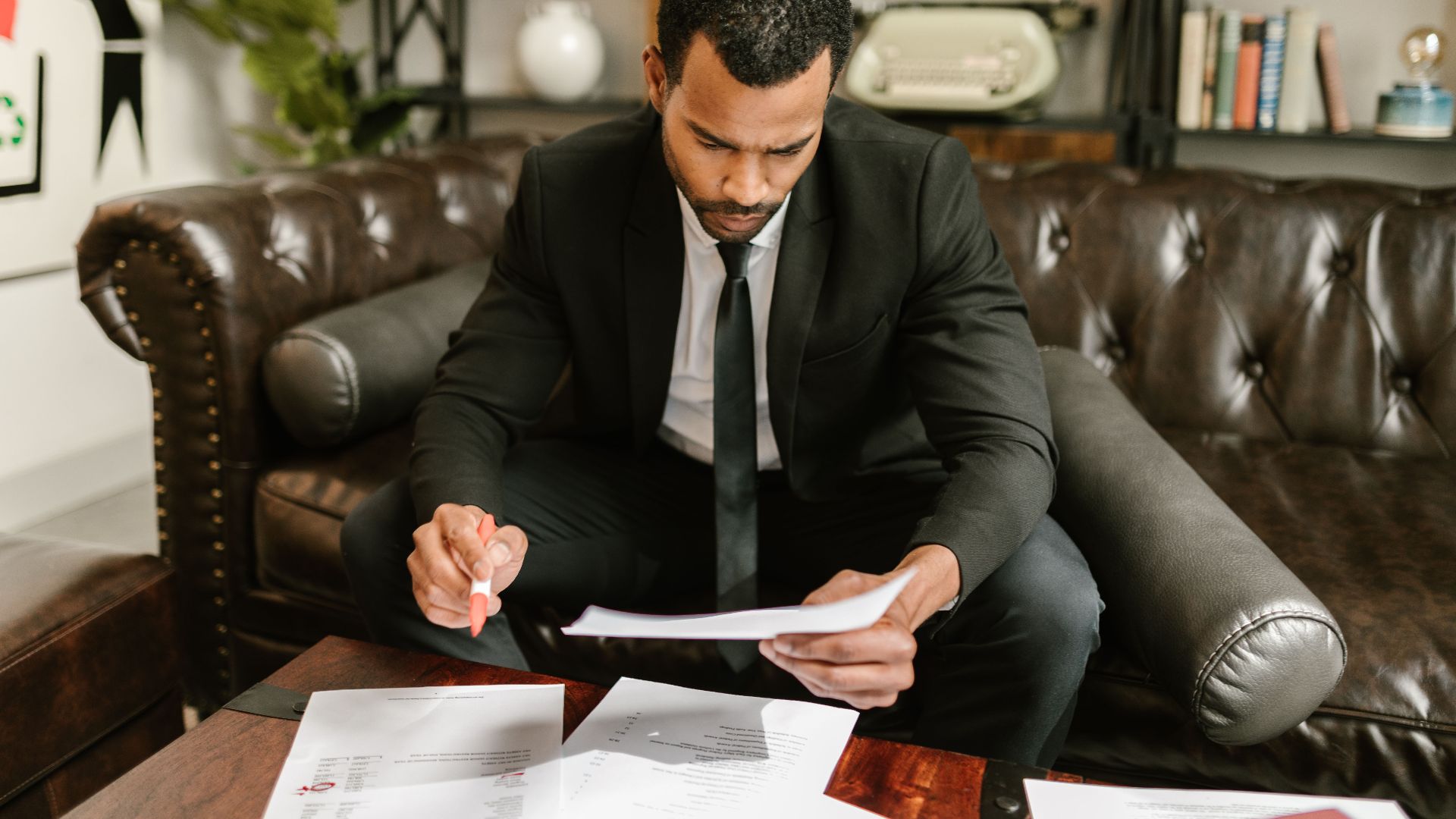 A businessman in a suit attentively reviews paperwork on a brown leather sofa.