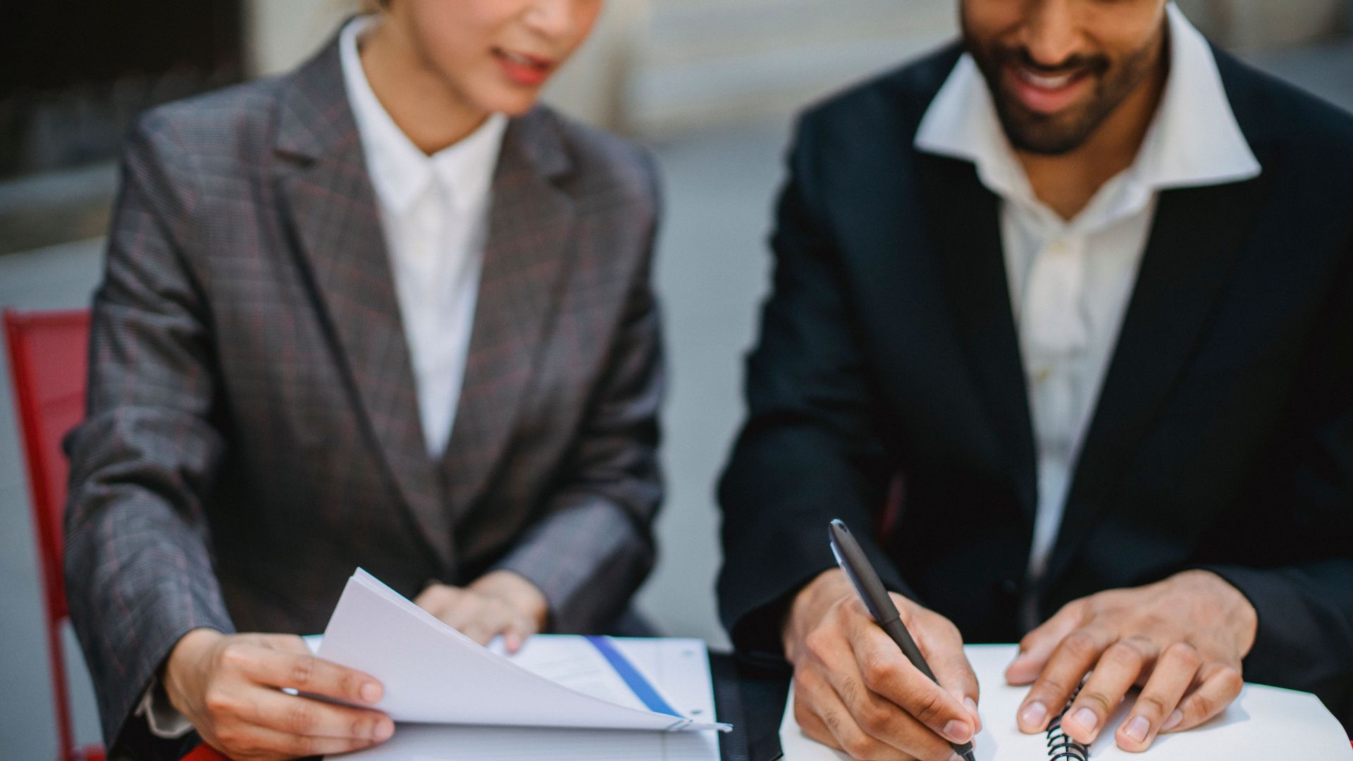 Two business professionals in suits reviewing documents and writing notes at a table.