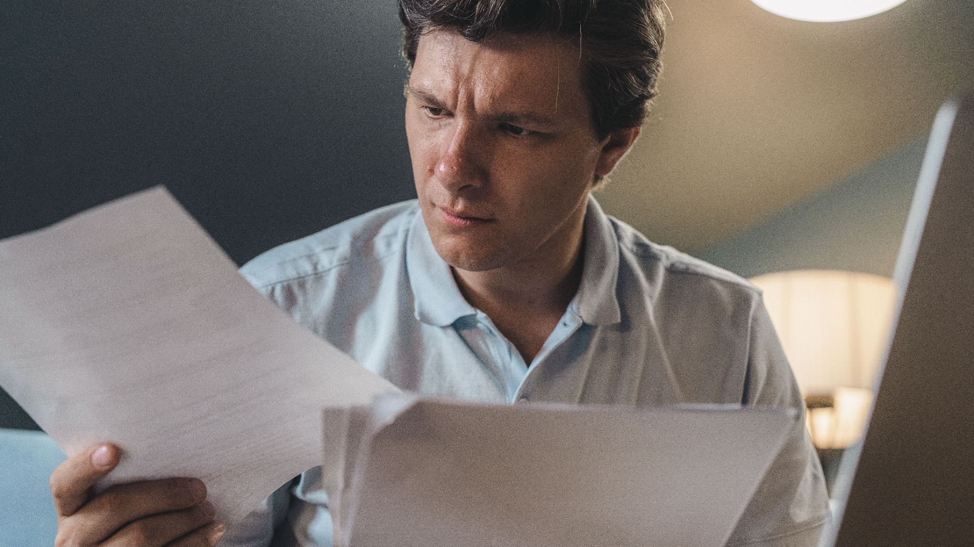 A man intently examines papers, seated indoors under warm lighting, focusing on his work.