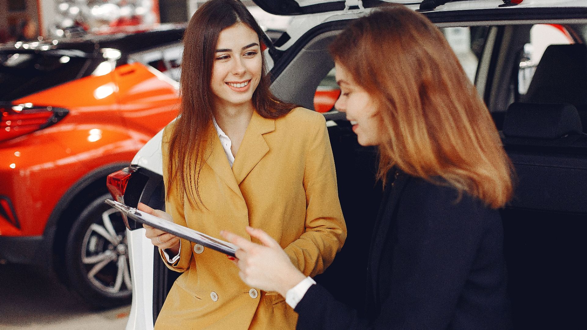 Positive smiling agent in trendy formal light brown suit discussing with happy excited female customer details of contract while leaning on car in showroom
