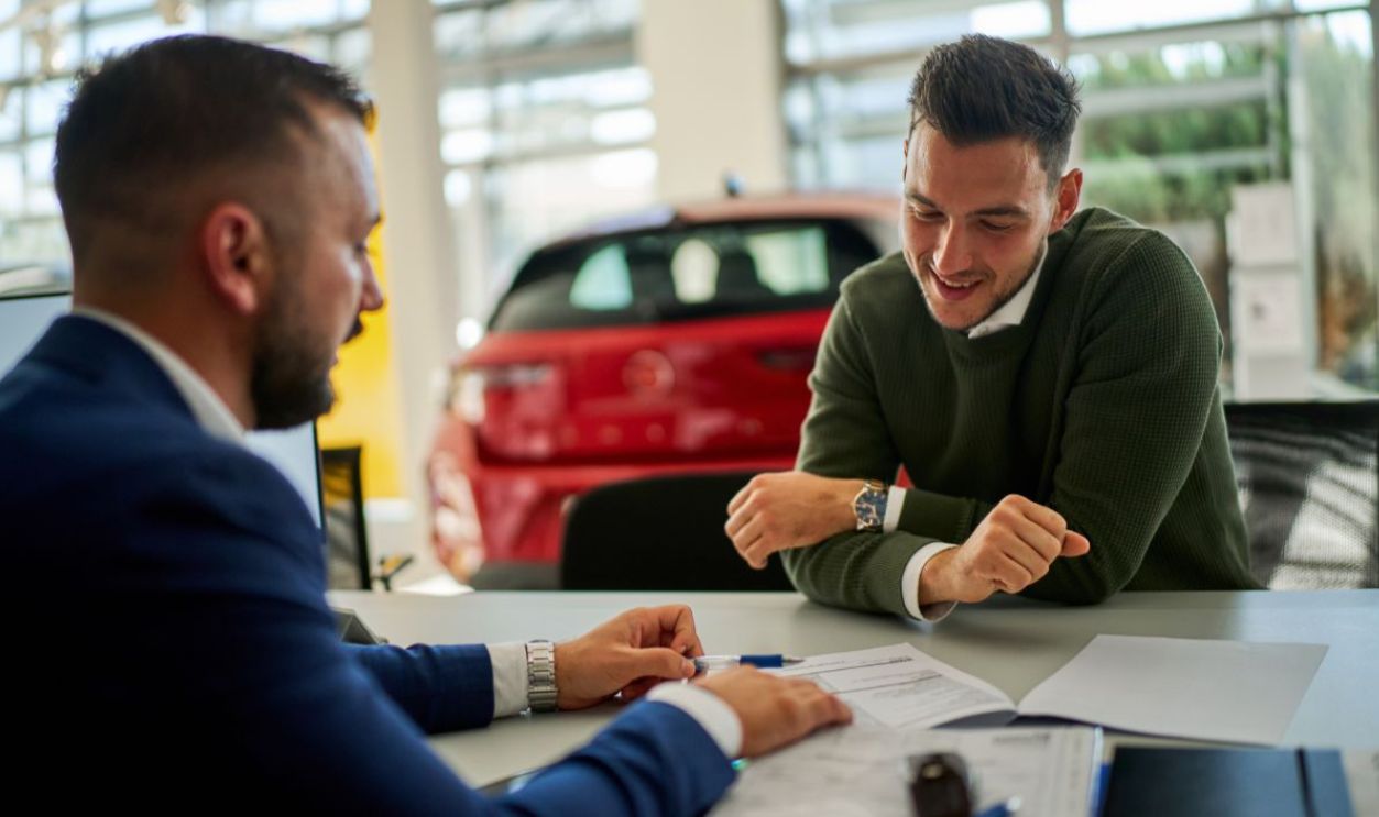 Car salesman and a customer engage in a discussion at a dealership table. Documents and a car key are on the table, with a vehicle visible in the background through large windows