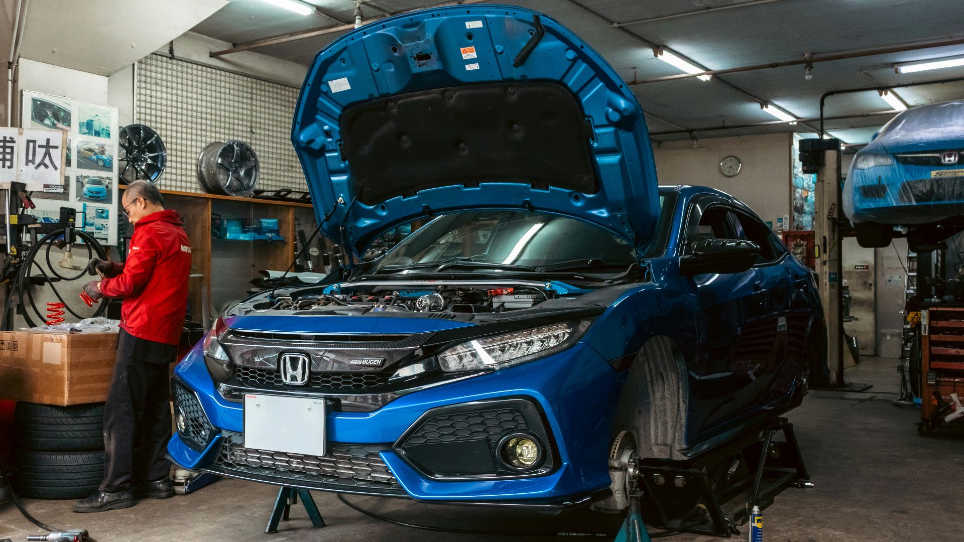 A mechanic in a red jacket working on a blue car in a garage