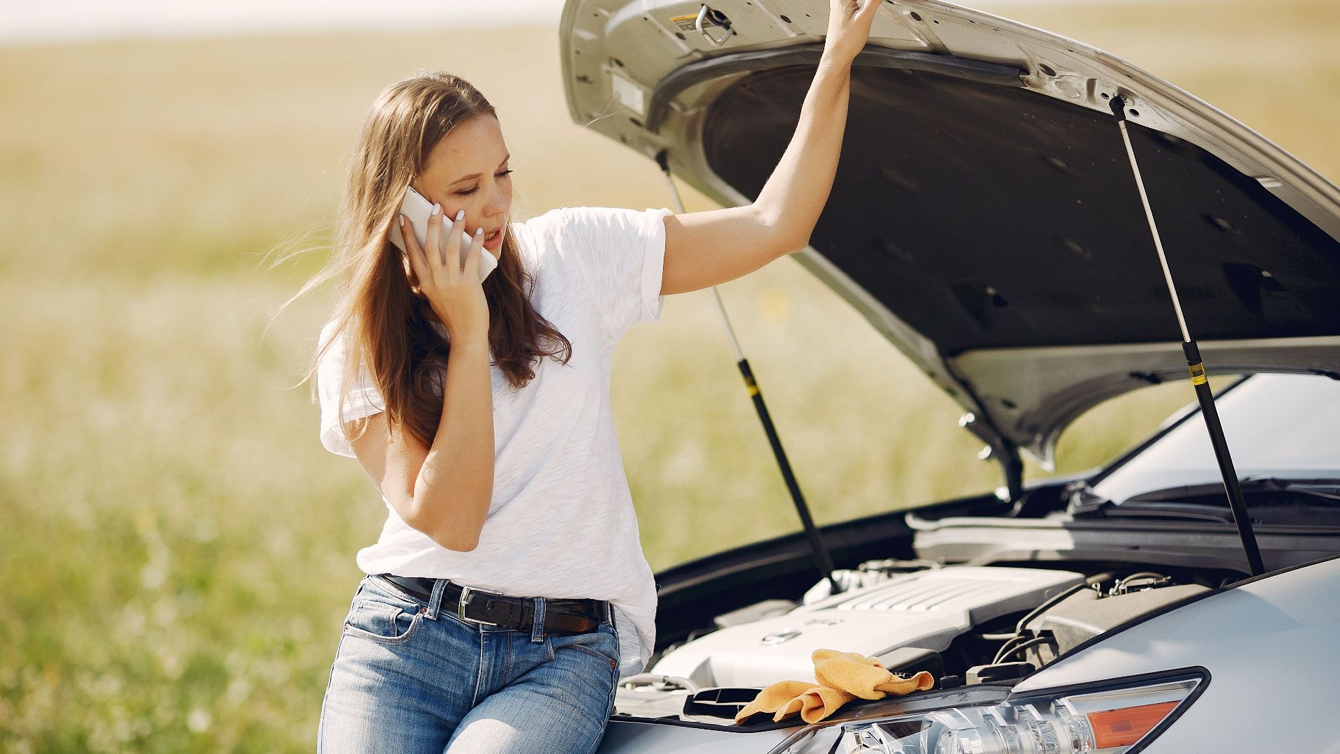 Worried young female driver in white t shirt and jeans speaking on phone while leaning on broken car with open hood asking for help with repair against green field in sunny summer day