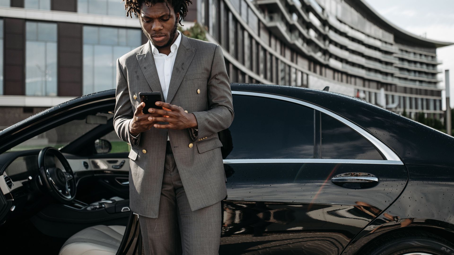 Professional man in a gray suit using his phone next to a luxury car.