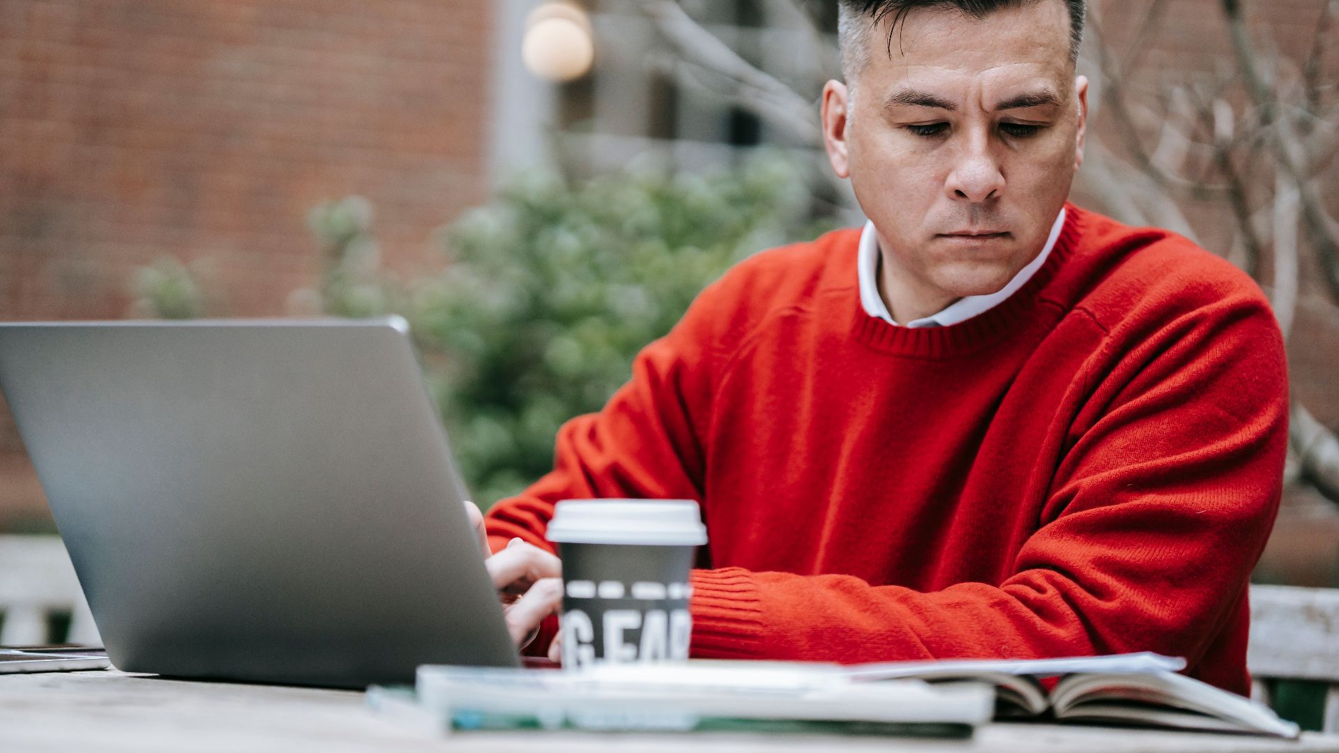 Adult man in a red sweater focusing on work on a laptop outdoors.