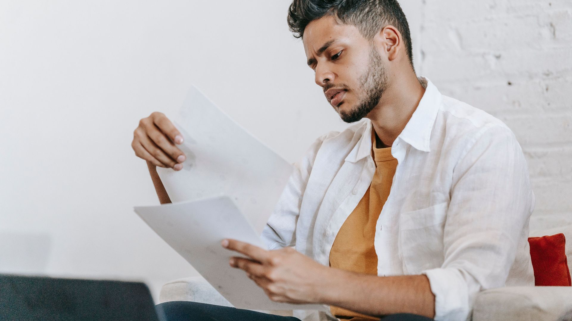 Concentrated ethnic male in casual wear reading documents and sitting on armchair near opened netbook