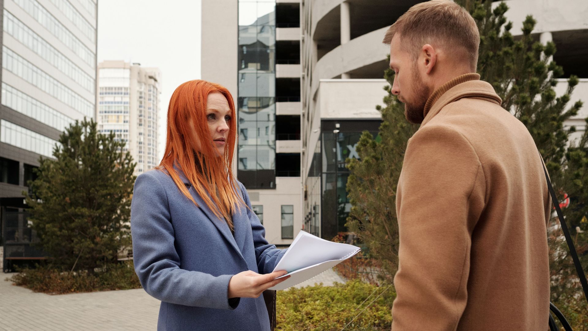Man and woman in corporate attire engaging in conversation outside urban office complex.