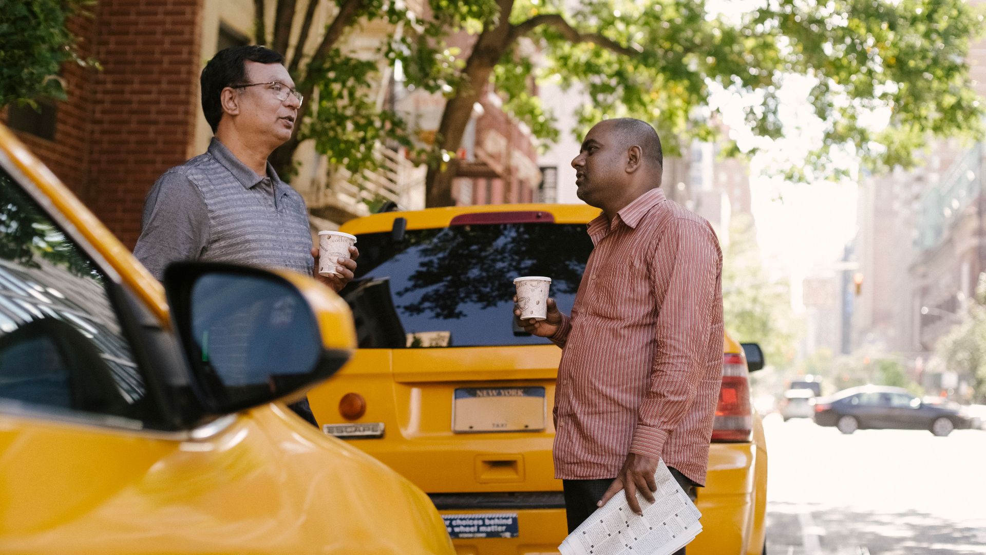 Side view of multiracial male coworkers with newspaper and takeaway coffee speaking near taxi autos while looking at each other in town