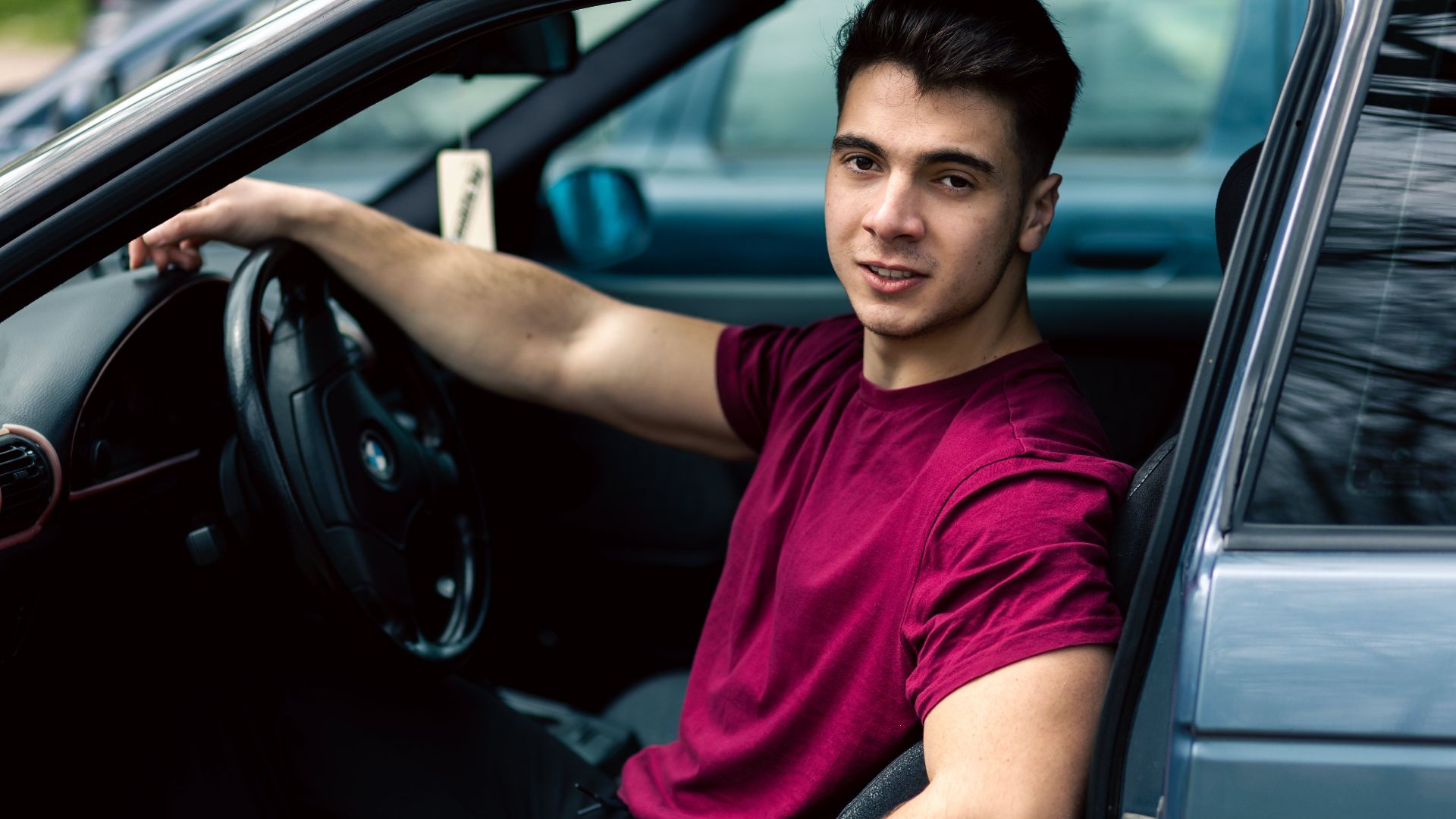 A young man wearing a maroon t-shirt sits casually in a blue car, portraying a relaxed and modern lifestyle.