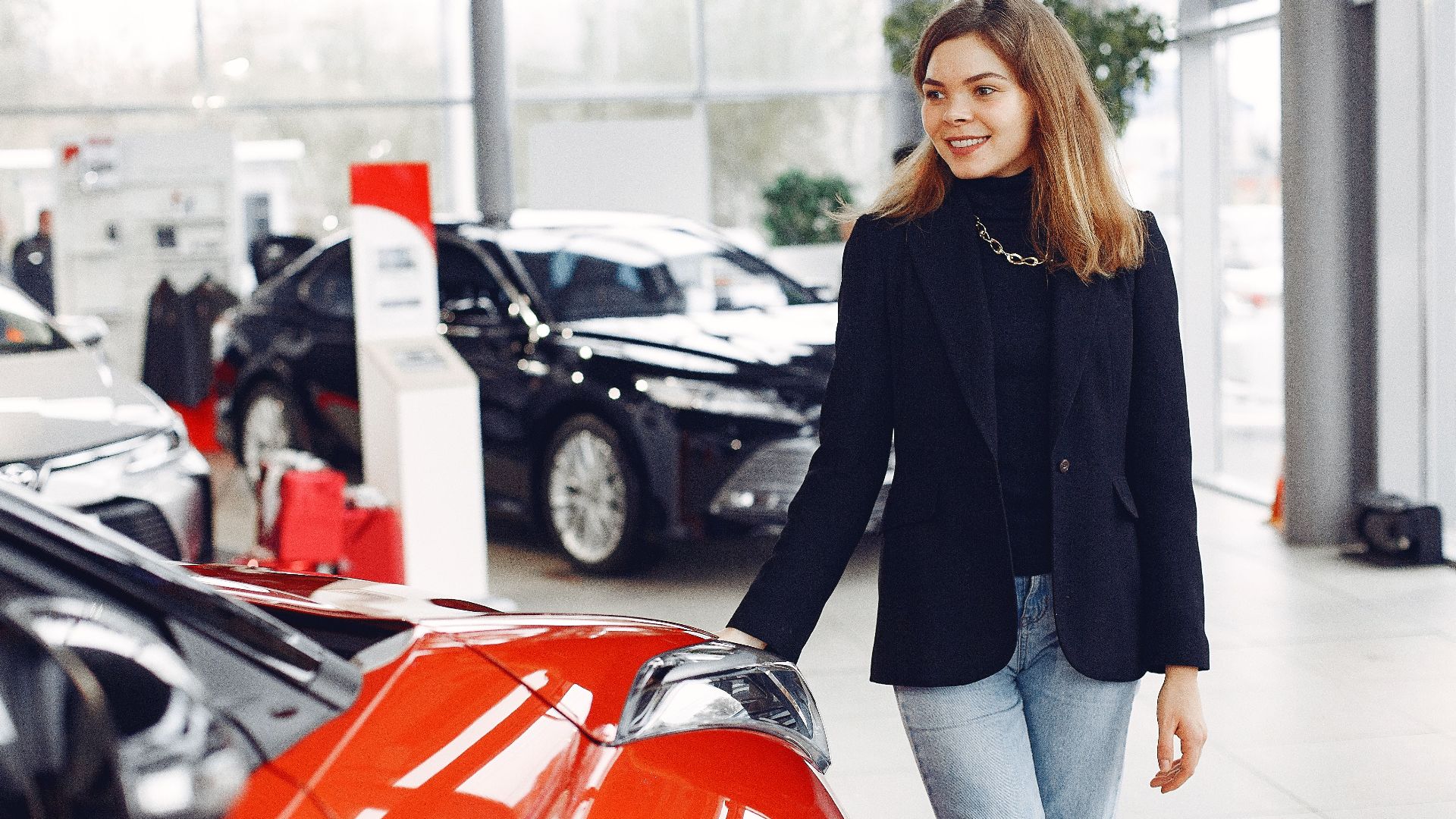 Happy stylish young female in jeans and black jacket standing near modern red shiny car in contemporary car showroom and looking at purchase dreamily