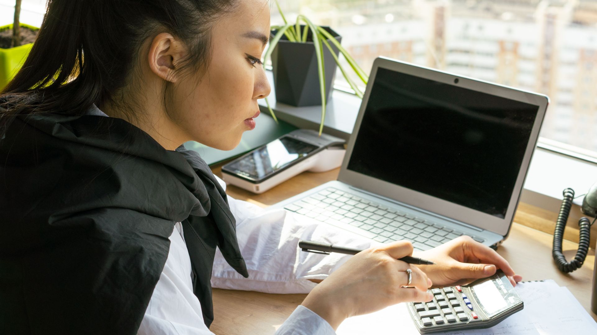 Asian woman working in a bright office, calculating finance on a desk with a laptop.