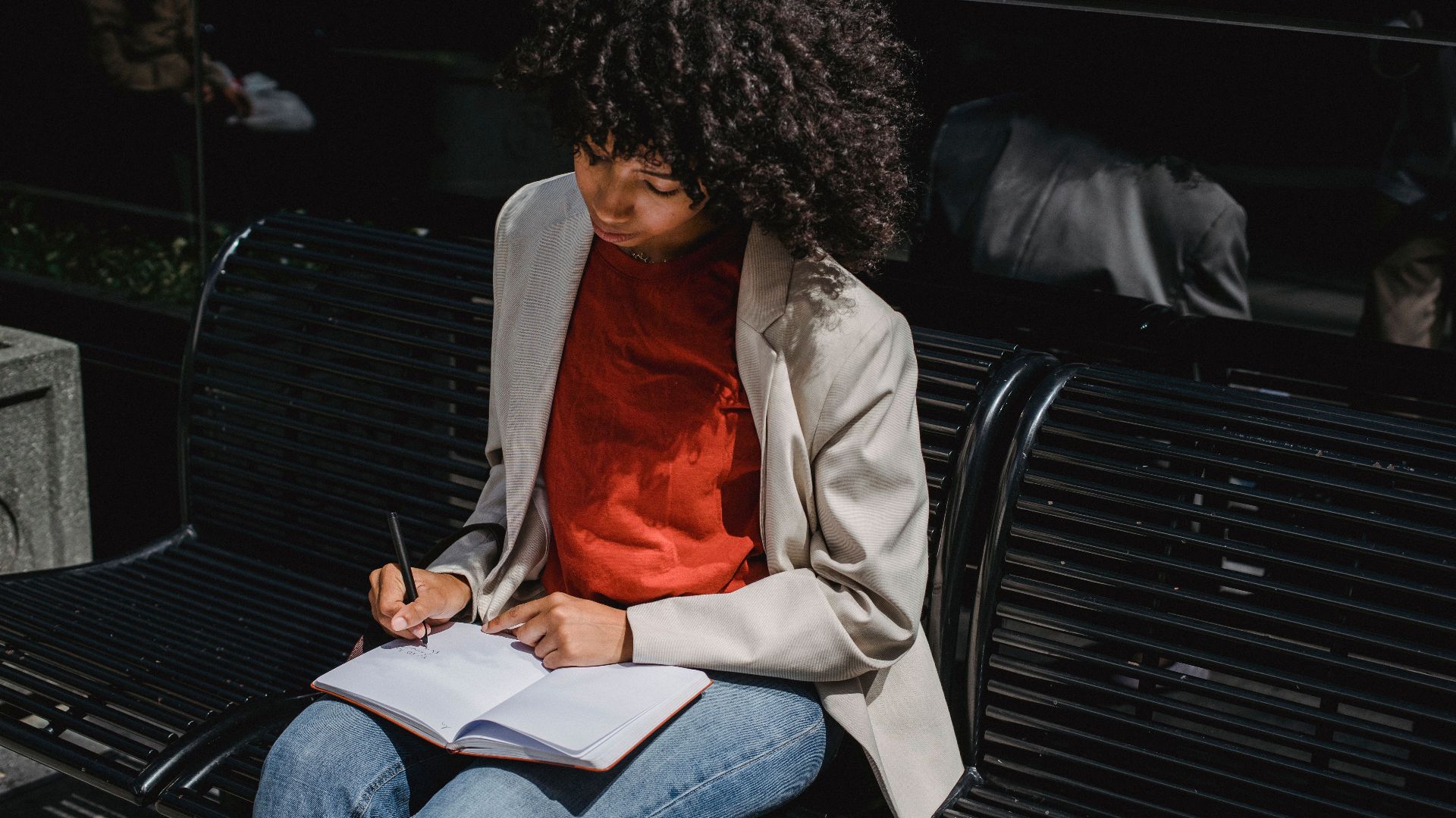 A young woman sits on a bench outdoors writing in a notebook during the day.