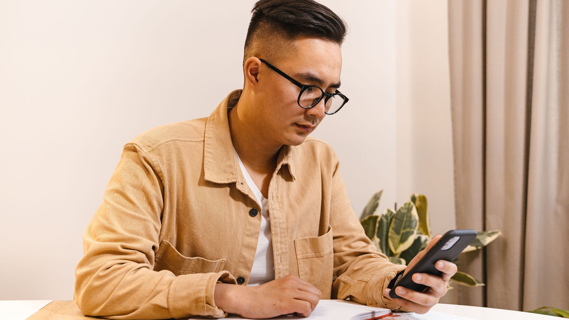 Adult male office worker concentrating on his smartphone and notebook at a desk indoors.