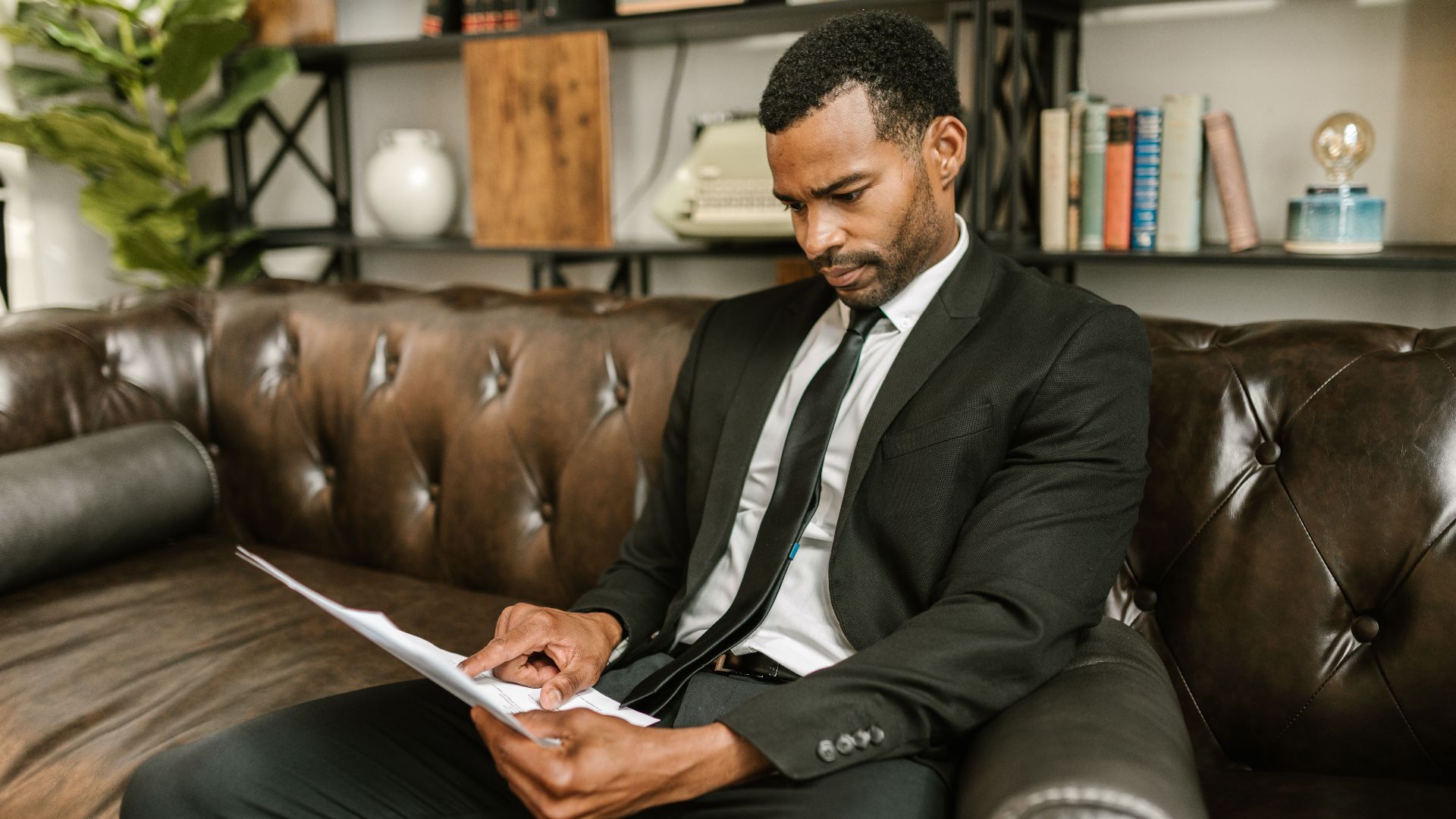 Businessman in corporate attire reading documents on a leather couch indoors.