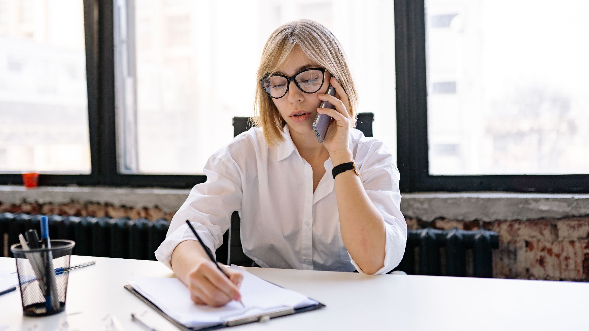 A woman in an office setting multitasking with a phone call and taking notes.