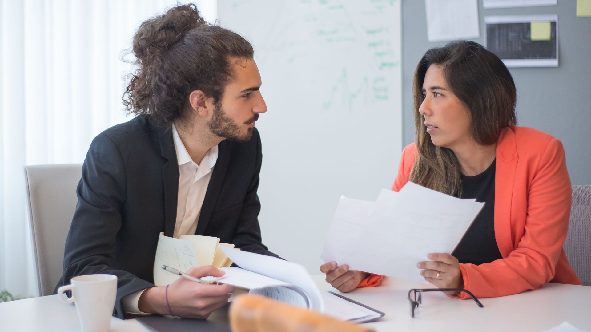 Two colleagues engaged in a discussion at an office table with documents and coffee.