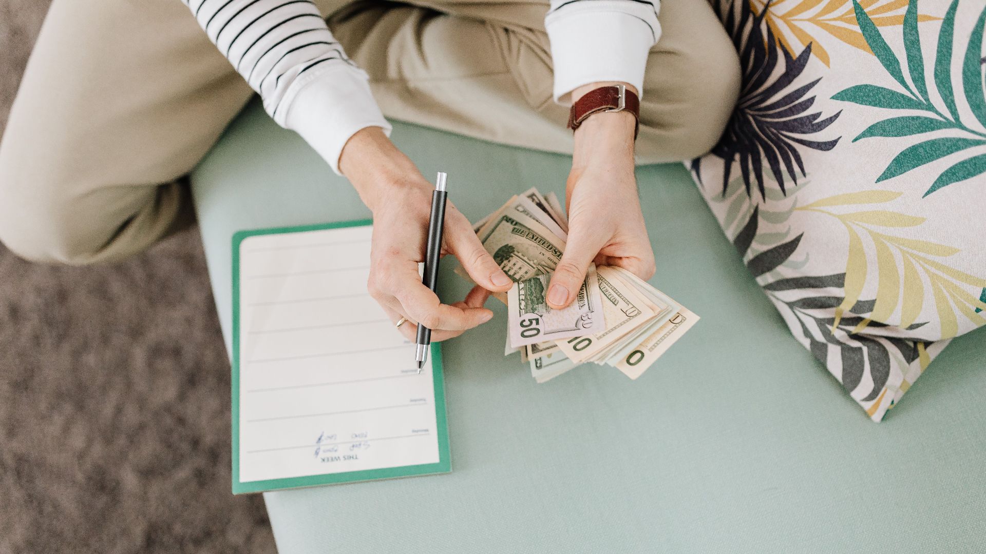 Person holding US dollar bills with a notepad and pen on a soft surface indoors.