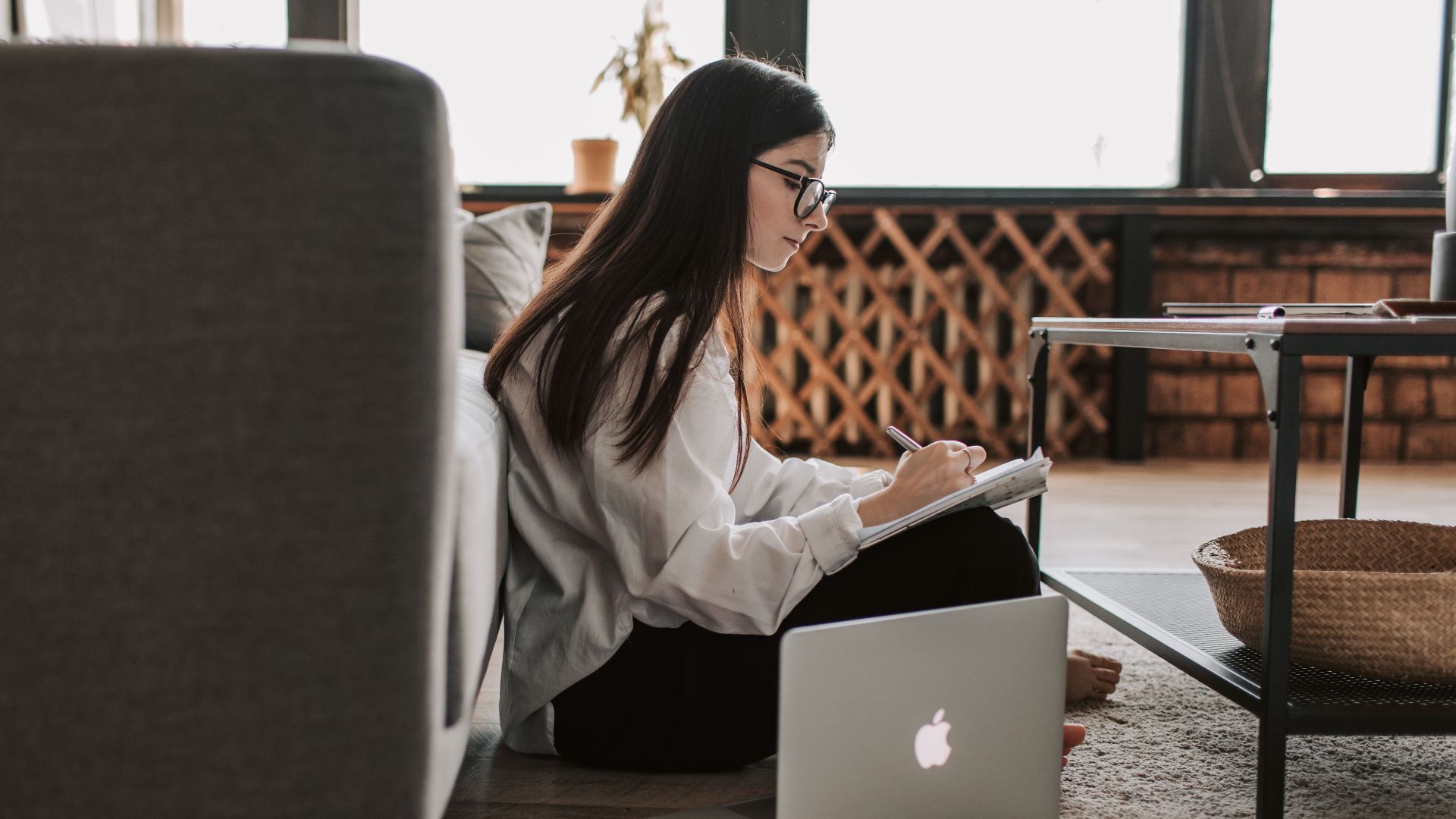 Woman sitting on floor with laptop and notebook, enjoying remote work in a comfortable home environment.