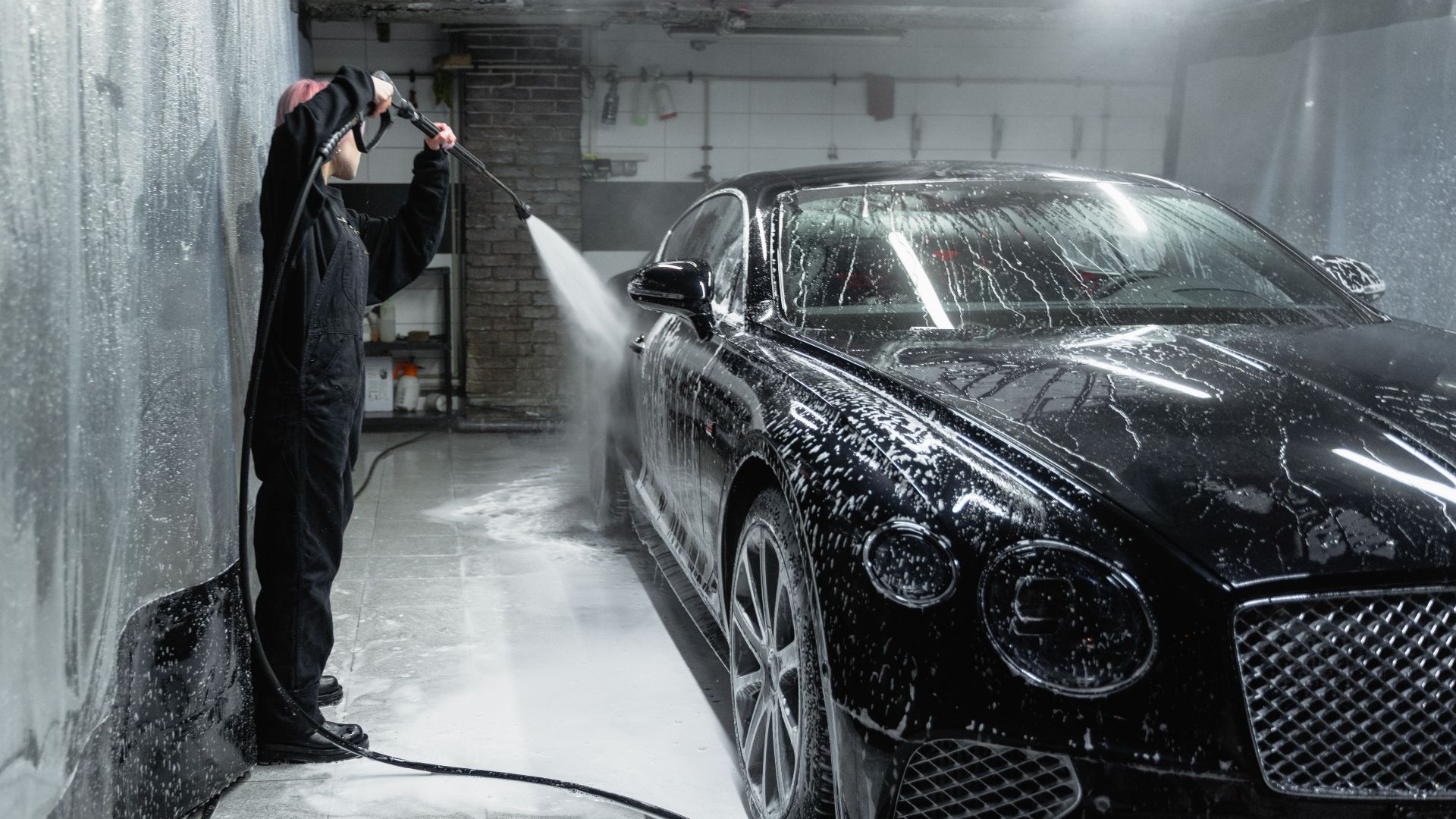 A person in a garage power washing a black car covered in soap and water spray.