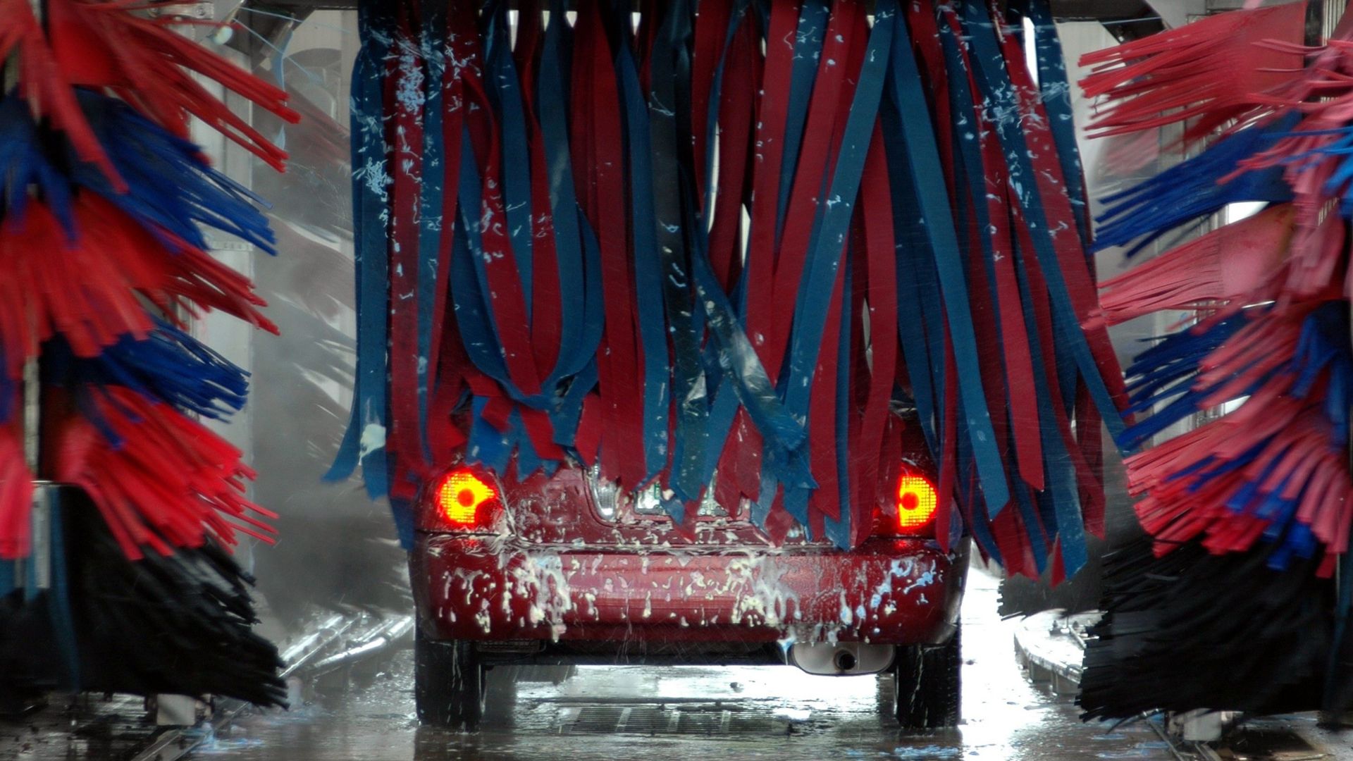 Red car being cleaned in an automated car wash with colorful brushes and soap suds.