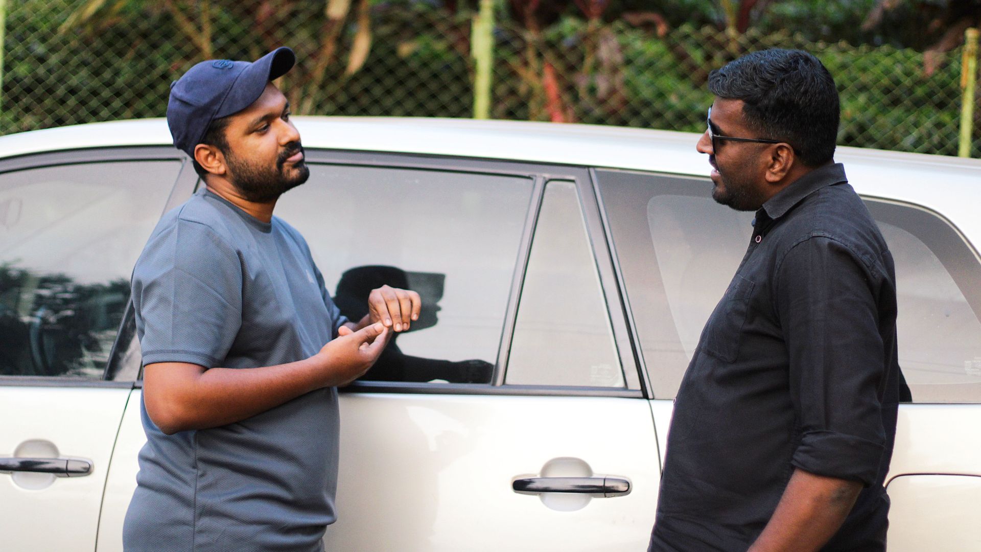 Two men having a casual discussion beside a car in an outdoor setting.