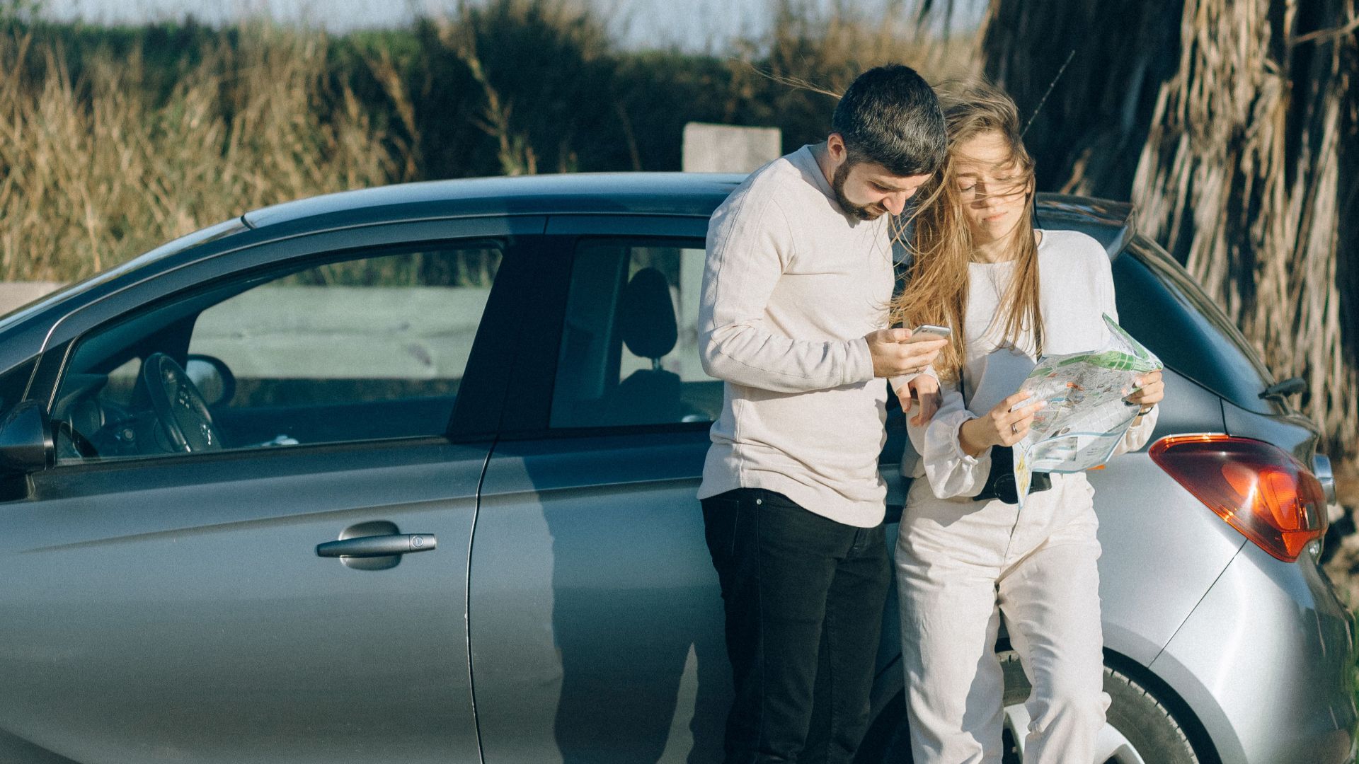 A couple standing by a car using a map to navigate during a sunny road trip.