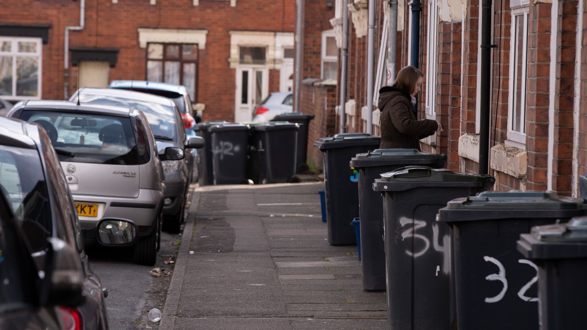 A street scene in a residential area with garbage bins and parked cars along the sidewalk.