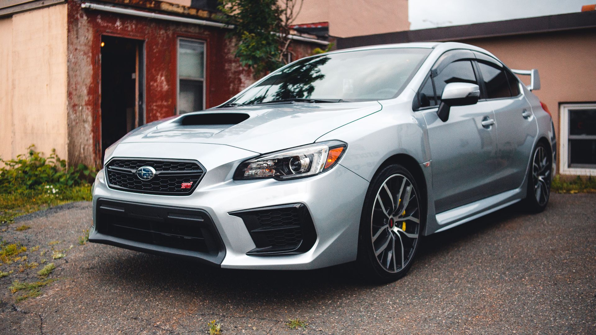 Silver Subaru WRX STI parked in a residential driveway with rustic backdrop.