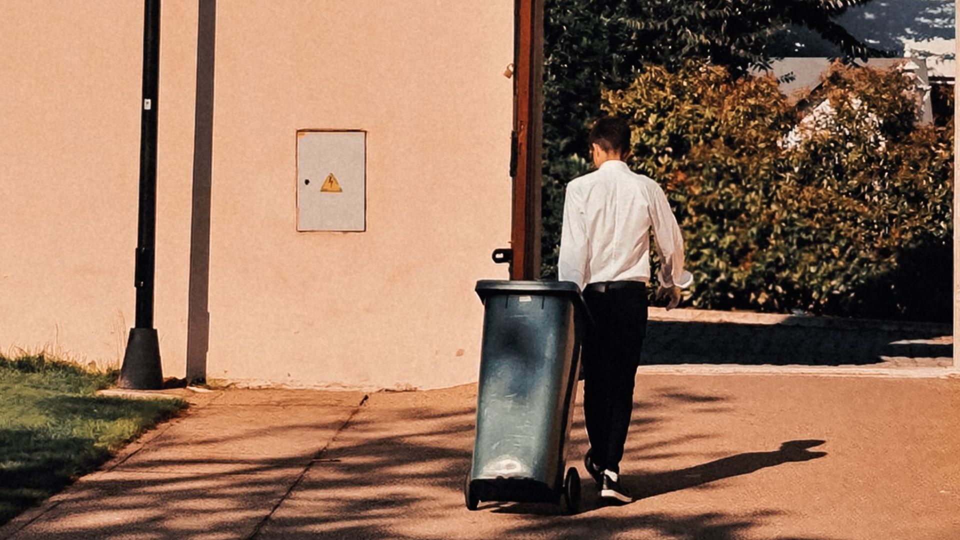 A person walks in a sunlit outdoor space, pushing a trash bin towards an arched exit.