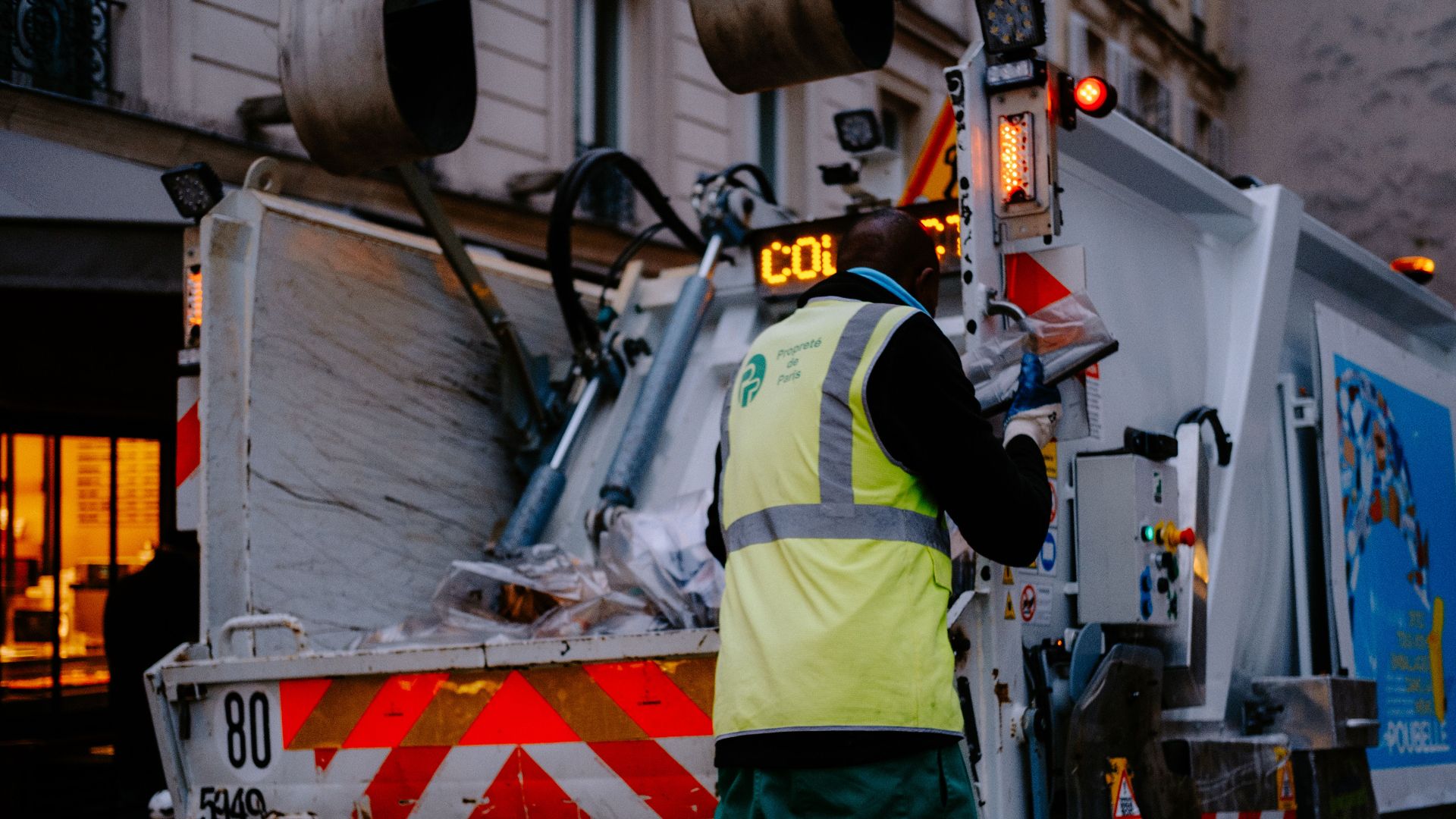A garbage collector works on a city street, managing waste collection at dusk.