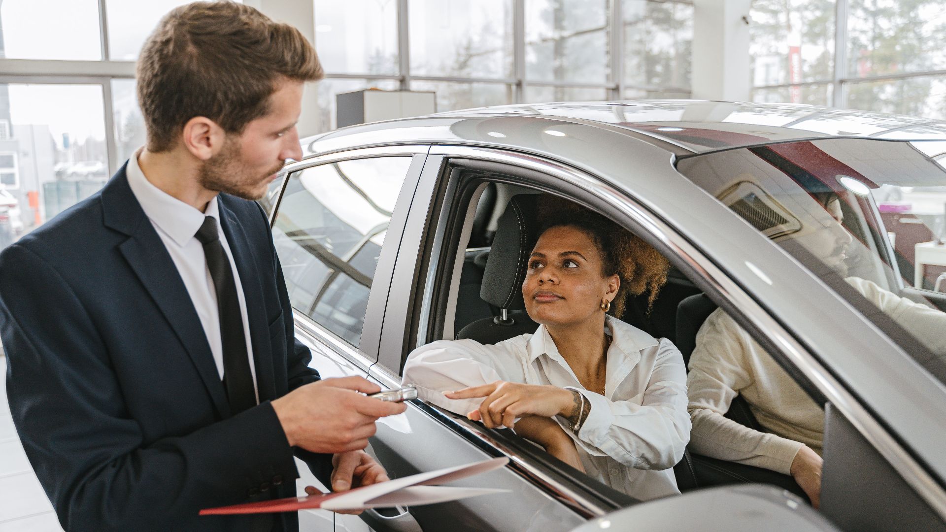 A car dealer hands keys to a woman sitting inside a vehicle in a car showroom.