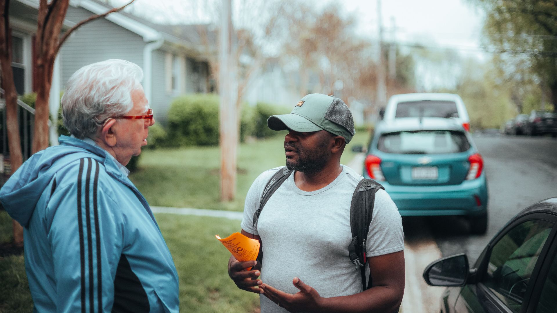 Two men engaged in conversation on a suburban sidewalk with parked cars.