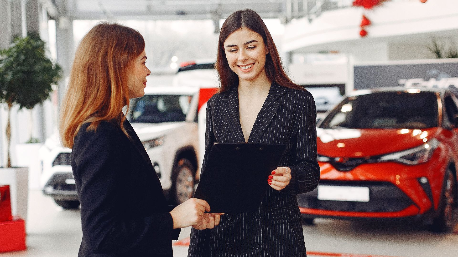 Cheerful young friendly dealer in formal stylish black dress showing contract to smiling female customer in black jacket while standing in car showroom against new shiny automobiles