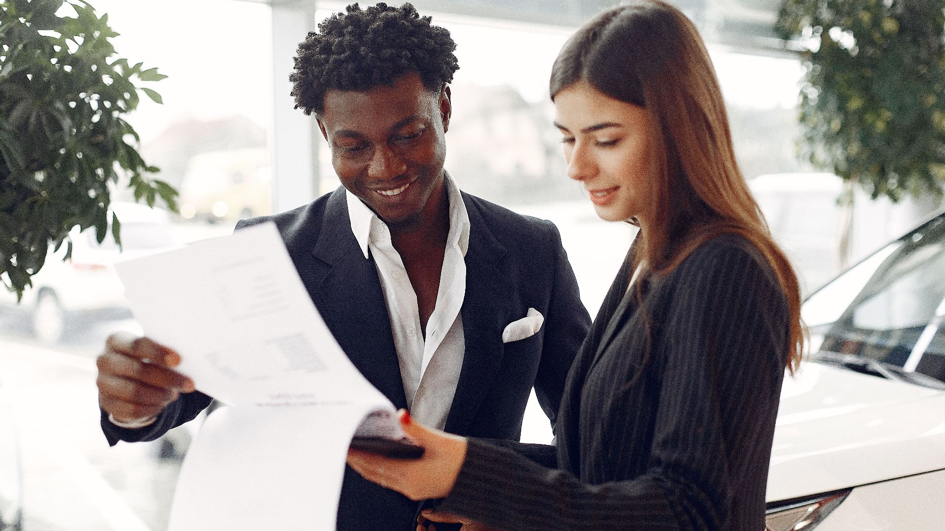 Cheerful smiling multiethnic businessman in classy suit and professional elegant female consultant standing close and reading contract details in car showroom
