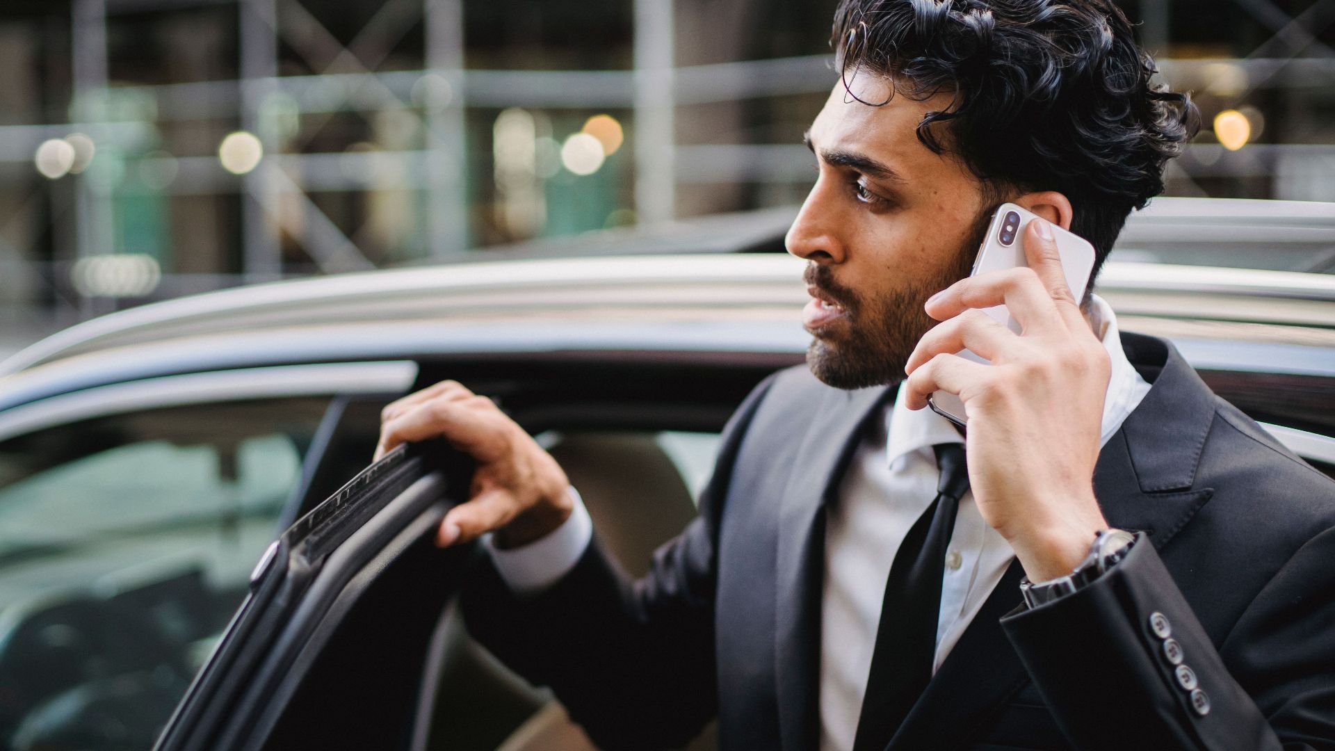 A businessman in a suit talking on his smartphone while exiting a vehicle.