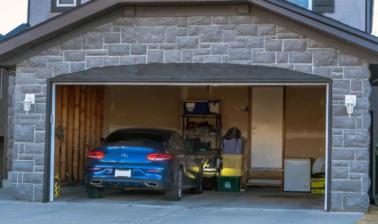 Calgary, Alberta, Canada. Nov 16, 2025. Open residential garage showing a blue Mercedes coupe and black sedan parked side-by-side amid storage shelves, lawnmower and concrete floor.
