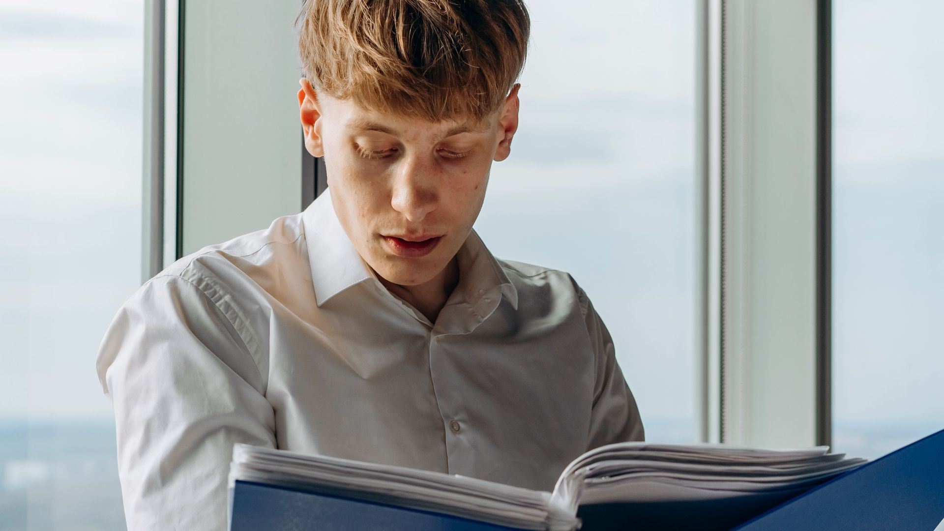 Businessman reading documents in an office with a city view, holding a blue binder.