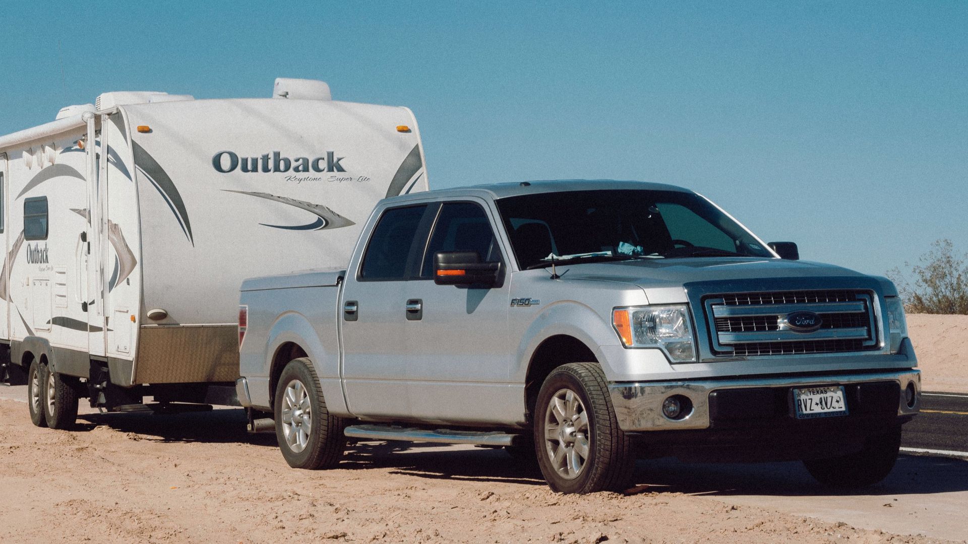 Silver truck towing an Outback trailer on a desert road under clear blue skies.