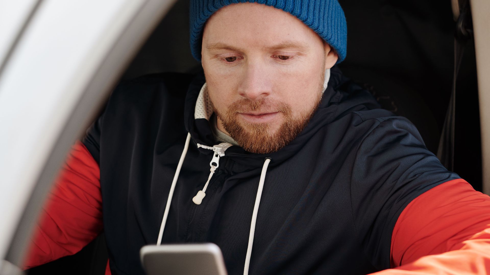 A man with a beanie uses a smartphone in a parked car.