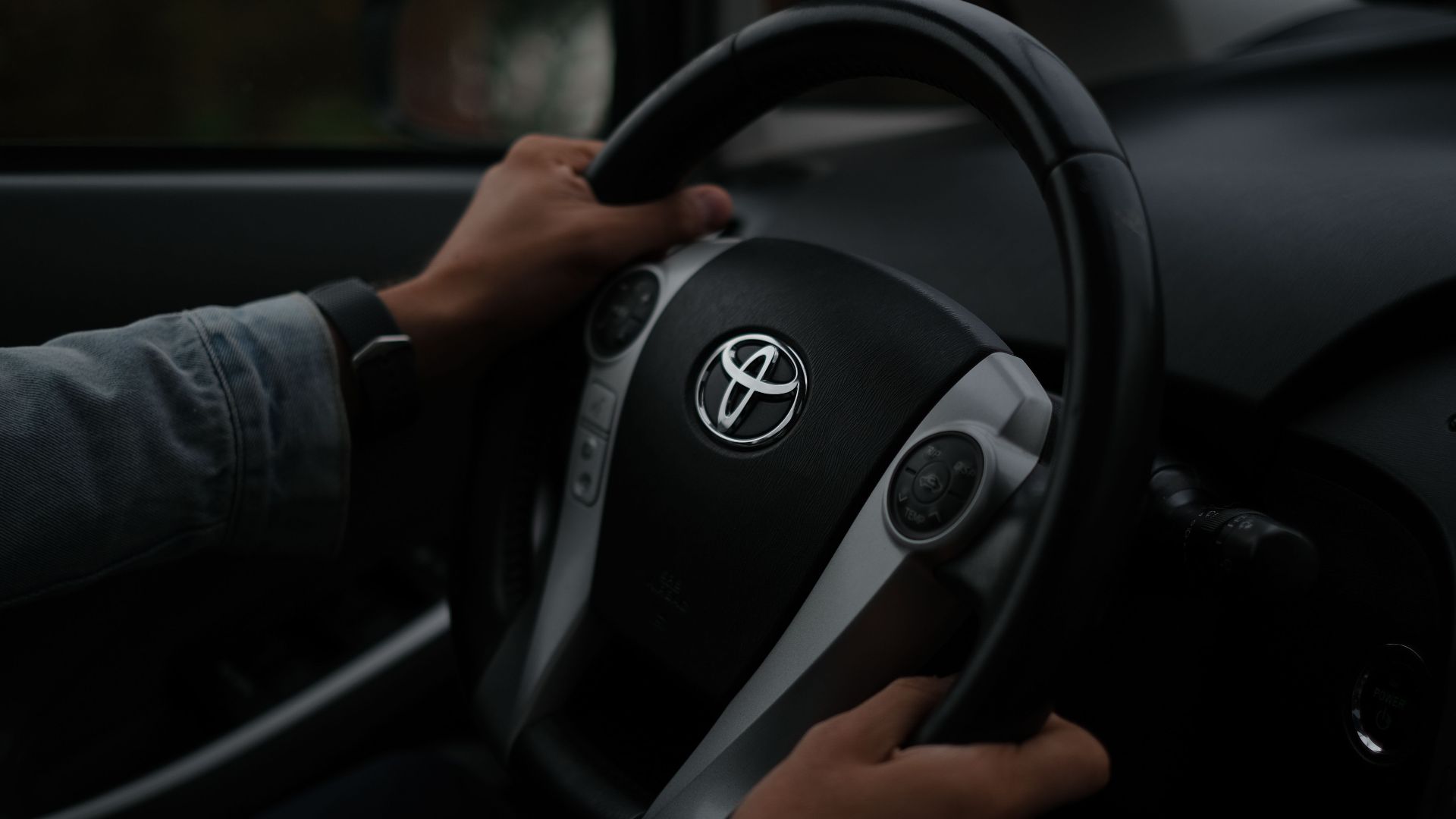 Hands on a Toyota steering wheel, closeup inside a vehicle. Darkened mood.