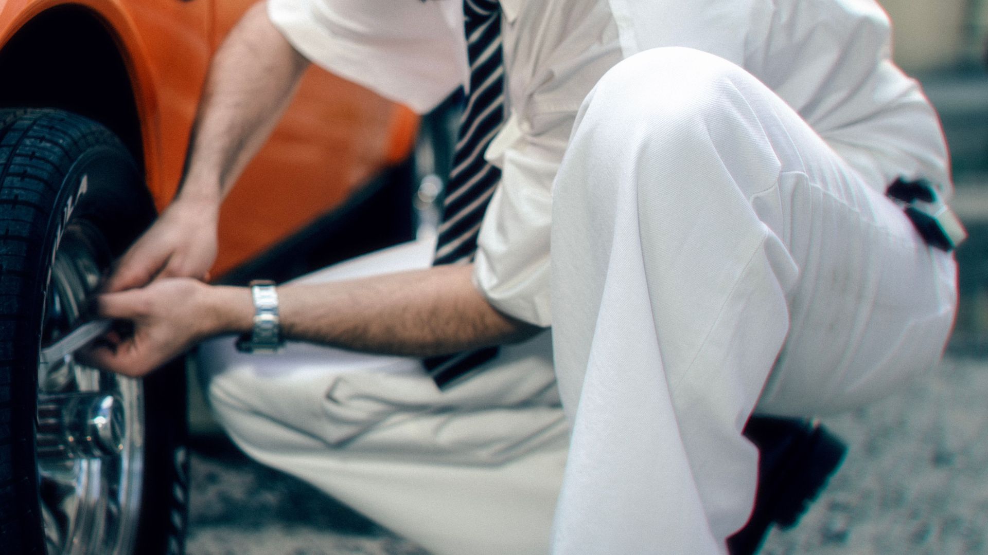 An adult man in formal attire kneels to fix a car tire on an urban street.