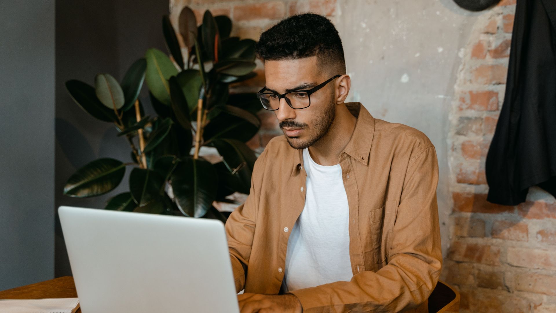 A man in glasses working on a laptop in a cozy cafe with brick walls and plants.