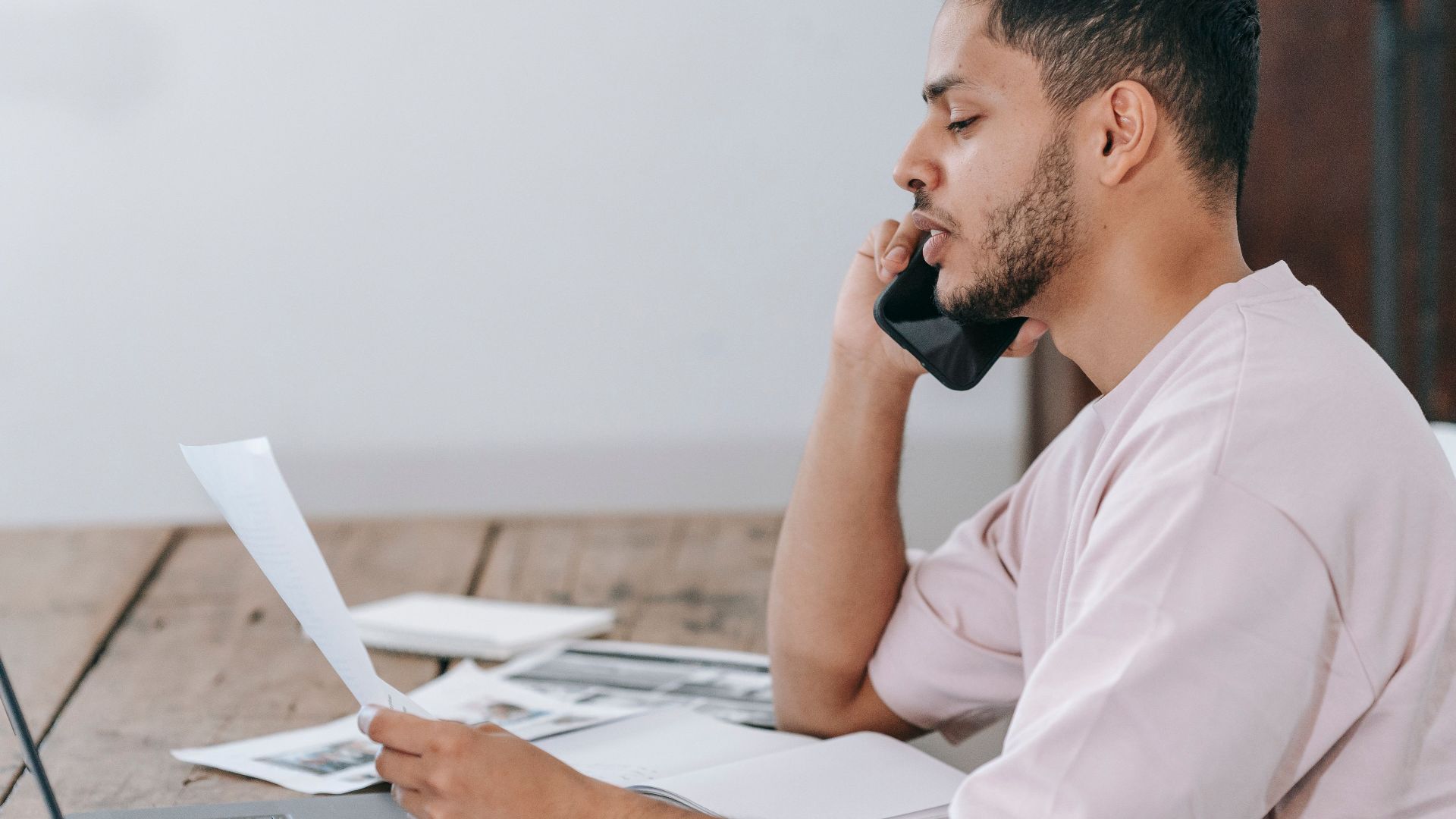 Side view of young Hispanic male remote employee discussing documents during phone call while working online with laptop and papers in home office