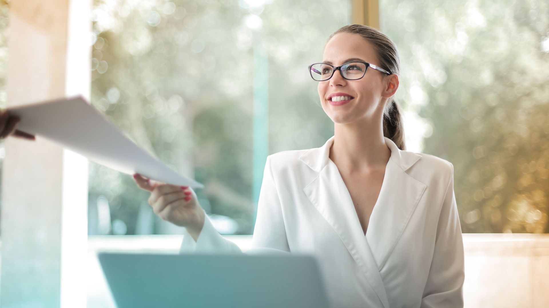 Low angle of successful female executive manager in classy style sitting at table with laptop in contemporary workplace and passing documents to colleague
