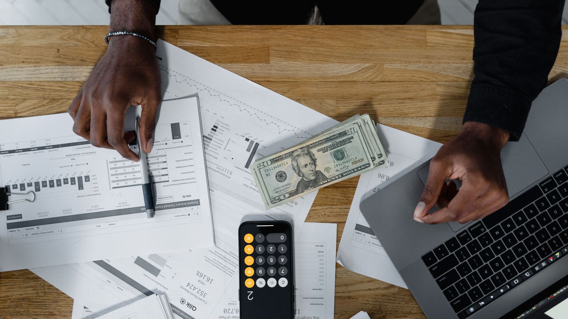 Hands calculate finances with papers, cash, and a laptop on a wooden desk.