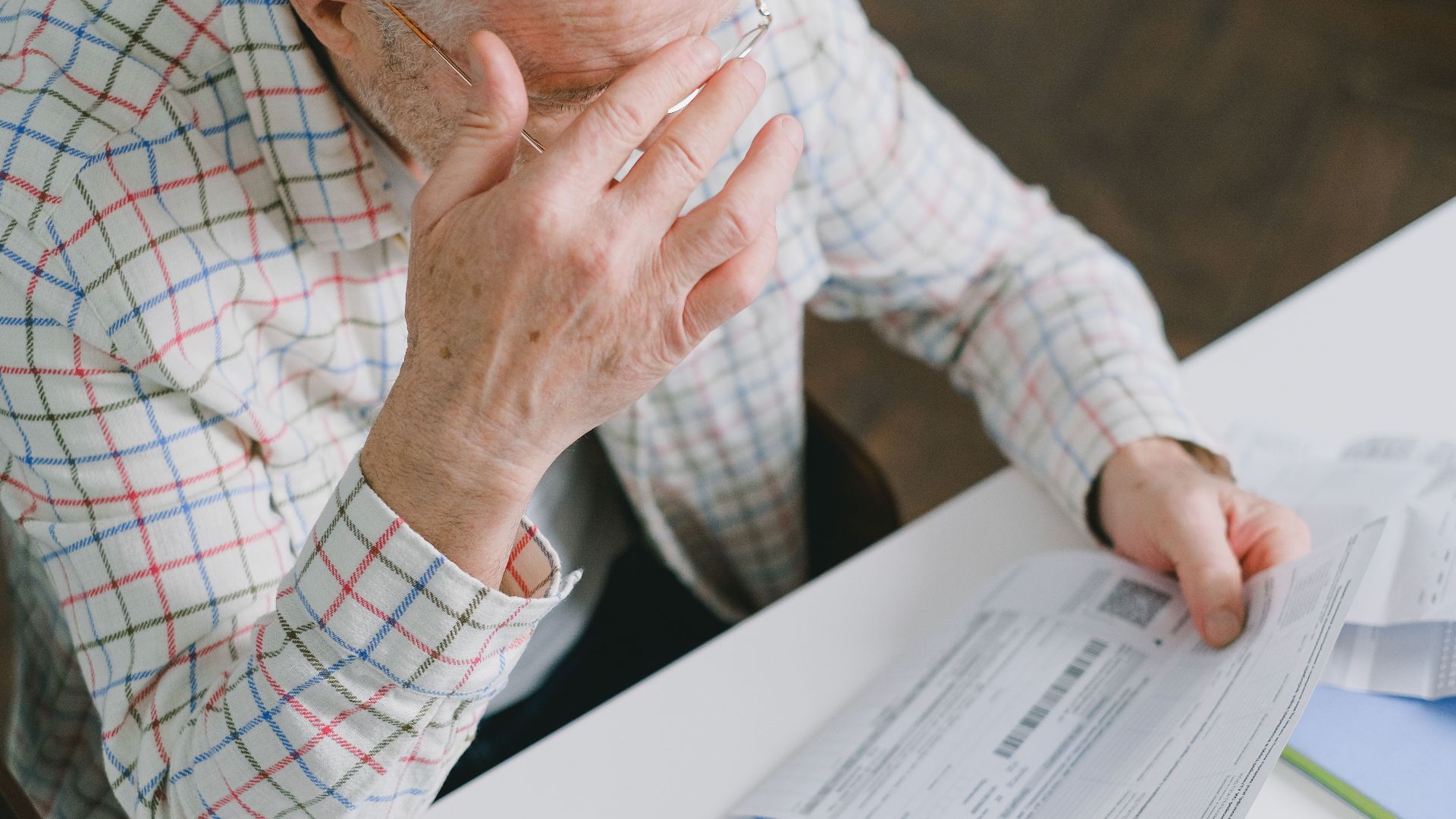 Elderly man in patterned shirt reading and holding bills at a home table, appearing focused.